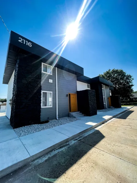 Apartment building at address 2116 under a bright sun. Gray siding, black stone columns, and vibrant yellow doors along a new concrete sidewalk.