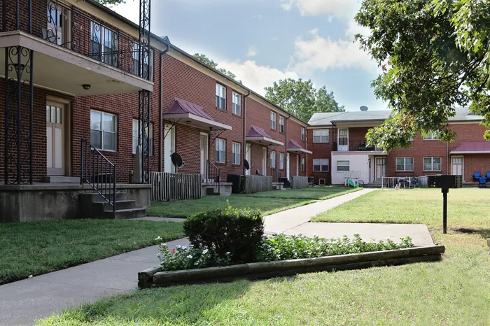 Two-story brick apartment buildings with small covered porches, decorative metal railings, and a shared grassy courtyard. A sidewalk runs through the courtyard, bordered by flower beds and trees under a partly cloudy sky.