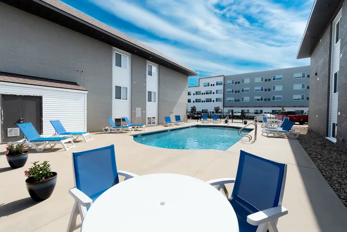 Outdoor pool area with lounge chairs and a round table with blue chairs under a bright sky. Surrounded by modern buildings, conveying a relaxing vibe.