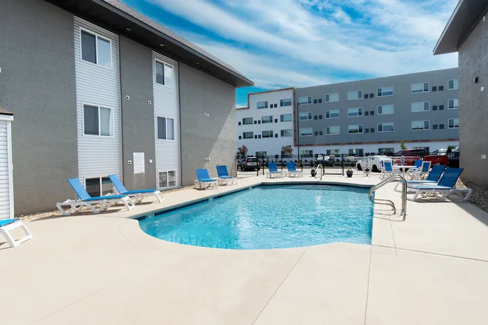 Modern apartment complex with a clear blue swimming pool, surrounded by lounge chairs. Buildings with large windows under a partly cloudy sky