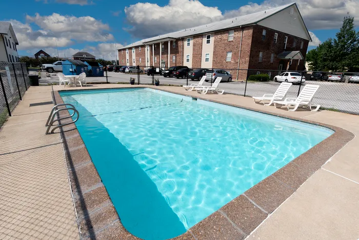 Outdoor swimming pool with clear blue water, surrounded by white lounge chairs on a sunny day. Brick building and parked cars in the background. Relaxing atmosphere.