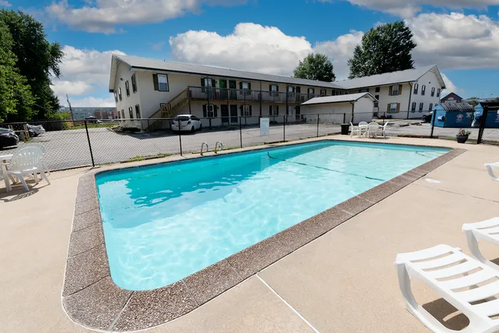 Outdoor pool at a motel with clear blue water under a partly cloudy sky. The motel's two-story building is in the background. Relaxed, sunny atmosphere.