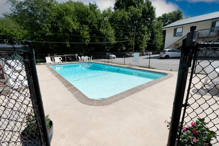 A serene outdoor swimming pool surrounded by a concrete deck, adjacent to a small parking lot and lush green trees under a partly cloudy sky.