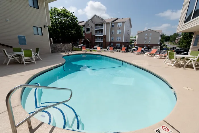 Swimming pool with surrounding lounge chairs &ndash; Curved outdoor swimming pool with steps, surrounded by tan pool deck, orange lounge chairs, and views of nearby apartment buildings at The Field Apartments.