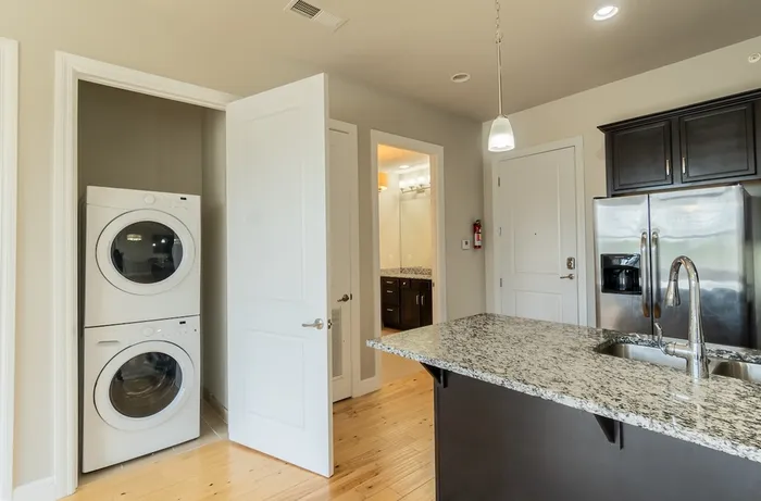 In-unit laundry at Chesterfield Lofts in Springfield, MO, featuring a stacked washer and dryer set within a convenient closet space. The laundry area is located near the kitchen, which includes dark wood cabinetry, stainless steel appliances, and a granite countertop, providing a seamless and functional apartment layout.