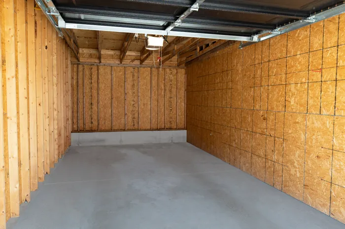 Interior view of a private rental garage at Chesterfield Lofts in Springfield, MO, featuring a spacious layout with unfinished plywood walls, exposed wooden beams, and a durable concrete floor. The open black roll-up garage door allows natural light to brighten the space, while an overhead light fixture provides additional illumination. This secure, enclosed garage offers residents a convenient storage and parking solution within this upscale Springfield, Missouri apartment community.