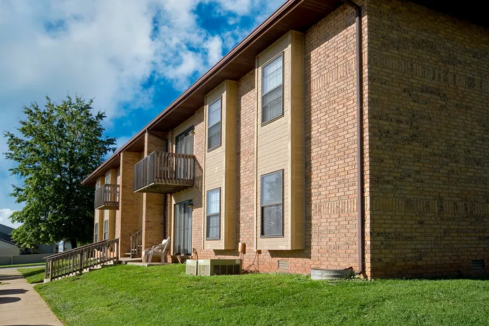 Exterior view of a two-story apartment building with tan siding and brick walls, small balconies on the upper level, and a sloping grass lawn under a blue sky with scattered clouds.