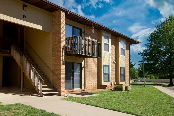 Exterior view of a two-story apartment building with brick walls and wooden paneling, featuring an outdoor staircase, a small wooden balcony, and a well-maintained grassy area under a clear blue sky.