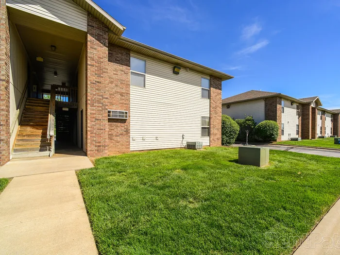 Side angle of a two-story apartment building with a neatly edged lawn, HVAC units, and a clear view of the open stairwell entrance.