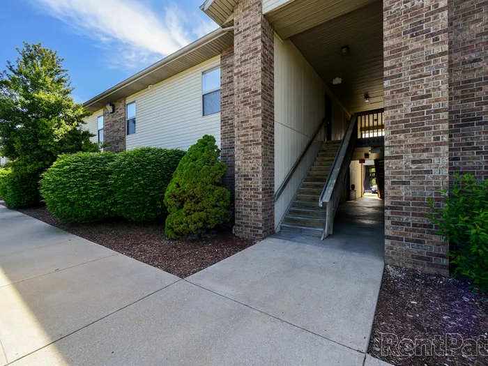 Front view of a Huntley Ridge East Apartments building featuring a central open stairwell, brick accents, and manicured bushes along the sidewalk.