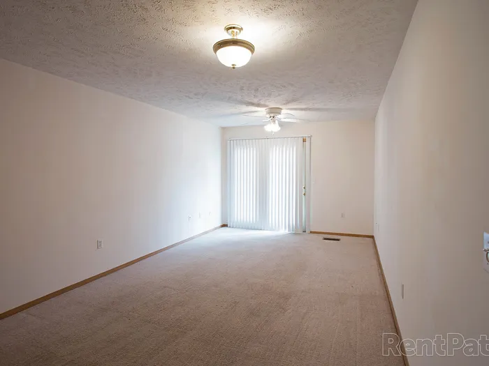 Spacious living room with carpeted flooring, ceiling lights, and a ceiling fan, leading to sliding glass doors with vertical blinds and a private patio or balcony.