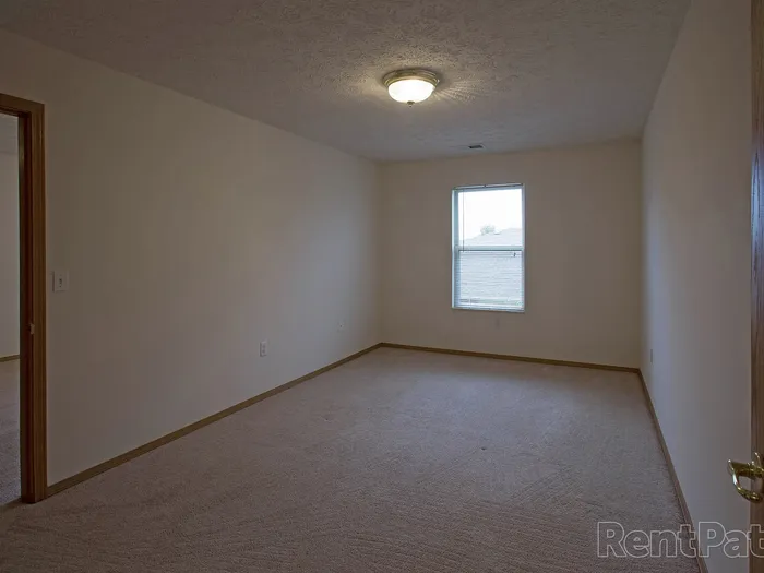 Carpeted living room at Huntley Ridge Apartments featuring a single window with blinds, neutral paint, and a spacious layout&mdash;ideal for flexible furniture arrangements.