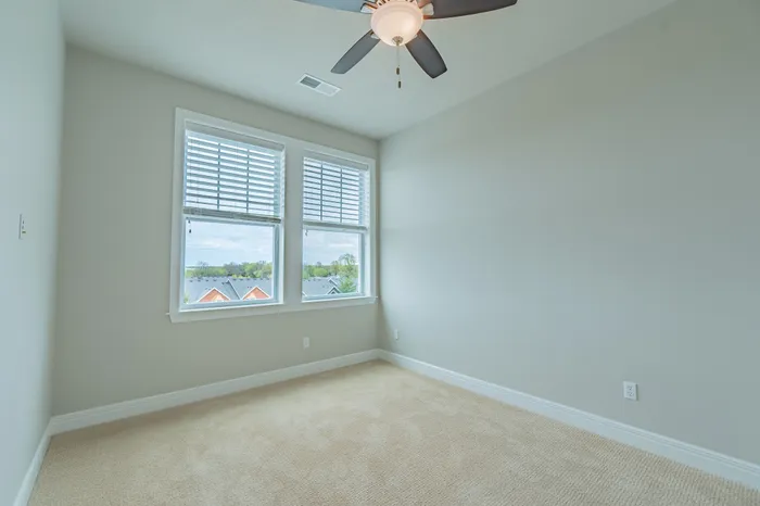 Bright bedroom at Chesterfield Lofts in Springfield, MO, featuring large windows with white blinds, soft neutral walls, and plush carpeting. A modern ceiling fan with a light fixture adds style and comfort, making this luxury apartment an inviting living space.