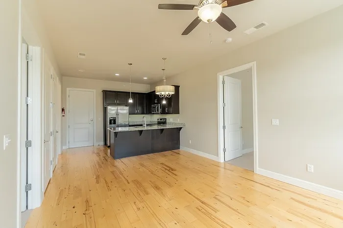 Spacious open-concept living area at Chesterfield Lofts in Springfield, MO, featuring natural hardwood flooring, neutral-toned walls, and a modern ceiling fan with a light fixture. The kitchen includes dark wood cabinetry, a granite island with a deep sink, stainless steel appliances, and pendant lighting. A doorway leads to a separate room, enhancing the functional layout of this luxury apartment.