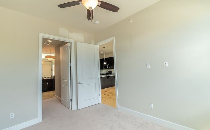Bedroom at Chesterfield Lofts featuring an open doorway leading to the kitchen and bathroom. The neutral wall tones, plush carpet, and ceiling fan enhance the cozy atmosphere