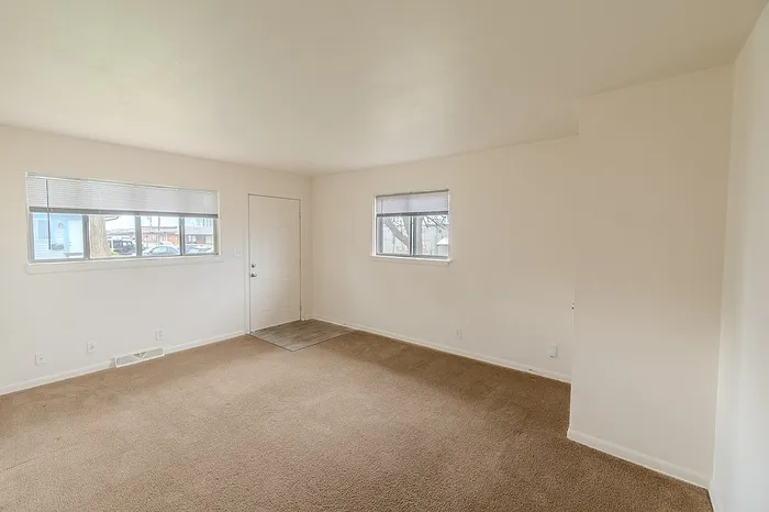 Living room at Village Club Apartments Springfield MO featuring beige carpet, off-white walls, and two windows allowing natural light. The front door and entryway with vinyl flooring are visible.