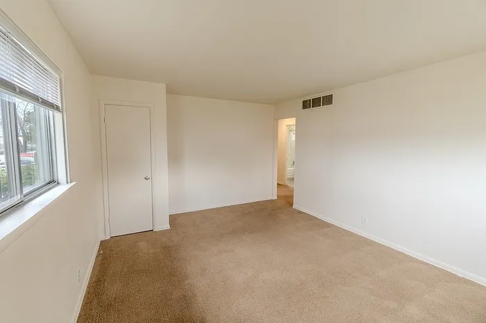 Open living space in a Village Club Apartment in Springfield MO with beige carpet, a window on the left wall, and a closet door. A hallway leads toward the bathroom and kitchen.