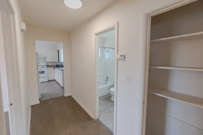 Hallway view in Village Club Apartments Springfield MO showing bathroom entry on the right, open shelving on the wall, and a kitchen with white cabinets and appliances straight ahead.