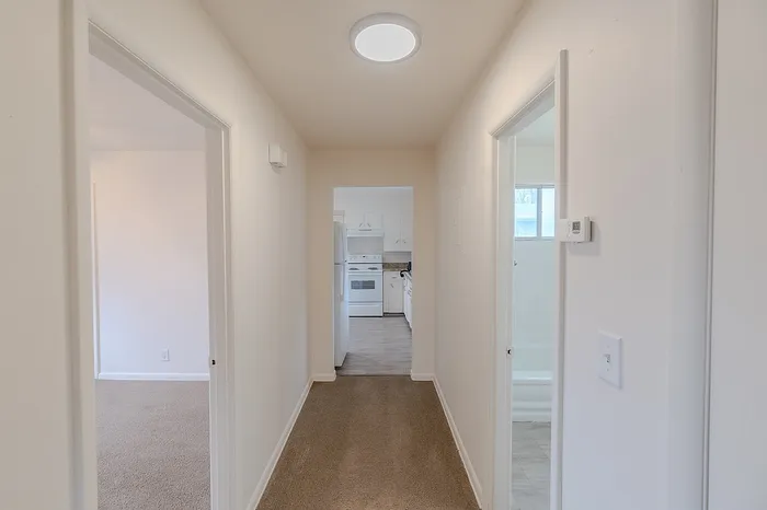 Narrow hallway inside Village Club Apartments Springfield MO with beige carpet, white walls, and a kitchen visible at the end. Doorways lead to bathroom and bedrooms.
