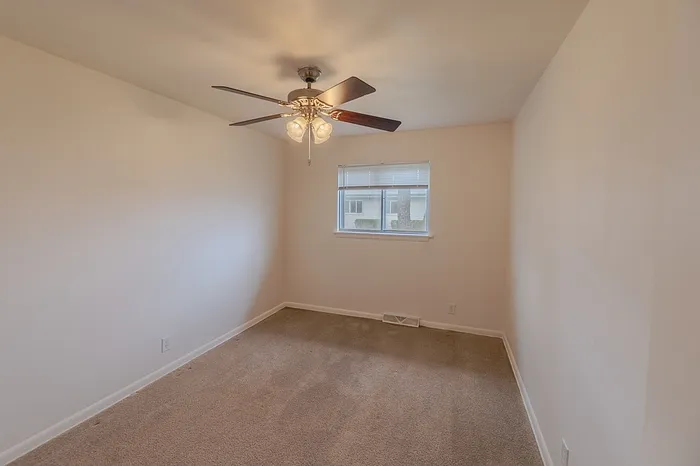 Bedroom at Village Club Apartments Springfield MO with beige carpet, white walls, a window centered on the far wall, and a ceiling fan with light.