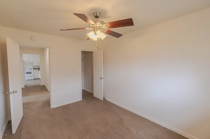 Bedroom in a Village Club Apartment in Springfield MO showing closet space with open door, beige carpet, and a ceiling fan with light. The hallway and kitchen are visible through the open bedroom door.