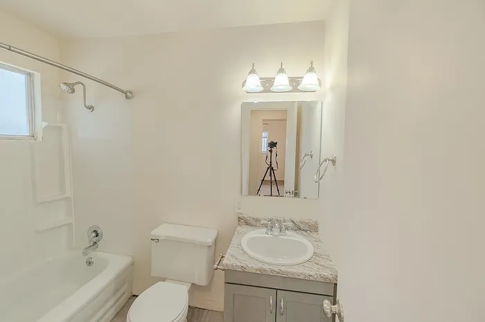 Full view of a bathroom in Village Club Apartments Springfield MO with a toilet, vanity with granite-look countertop, sink, mirror, and overhead lighting. A camera tripod is reflected in the mirror.