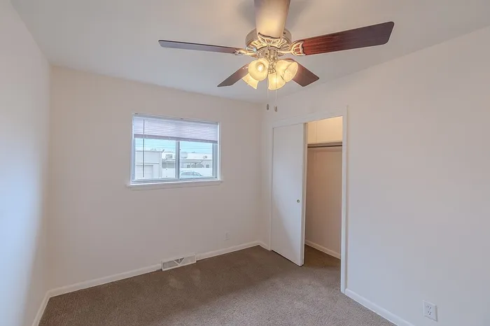 Bedroom at Village Club Apartments Springfield MO with a ceiling fan and light, sliding closet doors, beige carpet, and a window overlooking the exterior parking area.