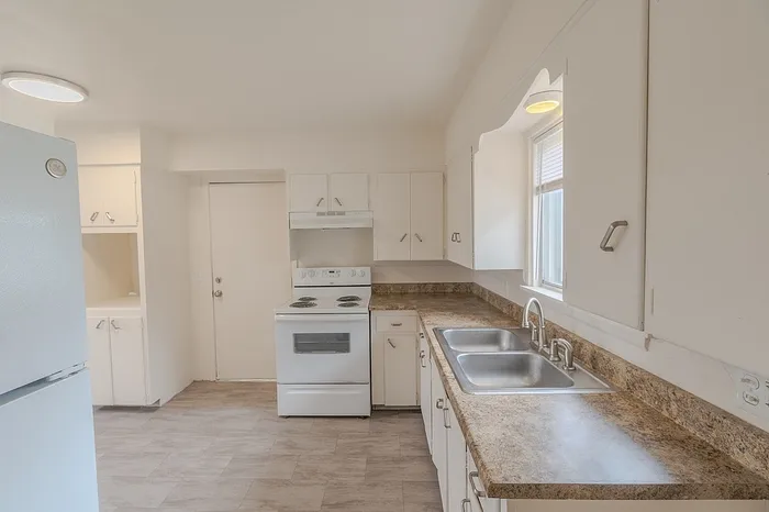 Kitchen at Village Club Apartments in Springfield MO featuring white cabinets, a white electric stove with vent hood, dual-basin sink under a window, and brown laminate countertops, offering practical layout for affordable housing residents.