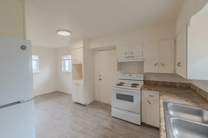 Wide-angle view of the kitchen at Village Club Apartments Springfield MO with white refrigerator, cabinets, stove, and dining nook near two windows, ideal for natural light and affordable living.