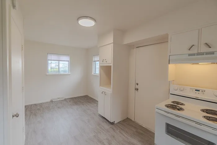 View from kitchen corner at Village Club Apartments Springfield MO showing white appliances, a door to the outside, and adjacent dining area with two windows, all with gray tile-look flooring.