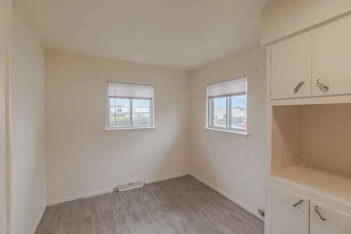 Corner of kitchen dining nook at Village Club Apartments Springfield MO with two bright windows, vinyl tile flooring, and a built-in upper and lower cabinet unit, designed for budget-conscious renters.