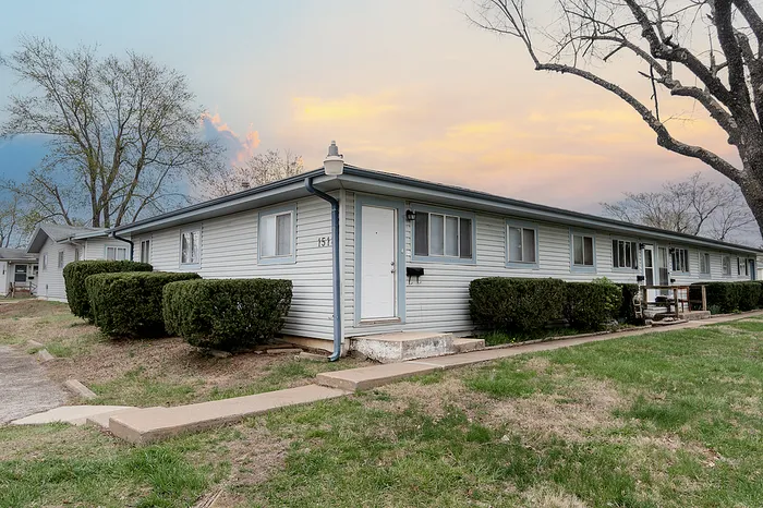 Exterior corner view of Village Club Apartments in Springfield MO, showing a single-story building with light siding, trimmed bushes, and concrete steps leading to a front door under a pastel sunset sky.