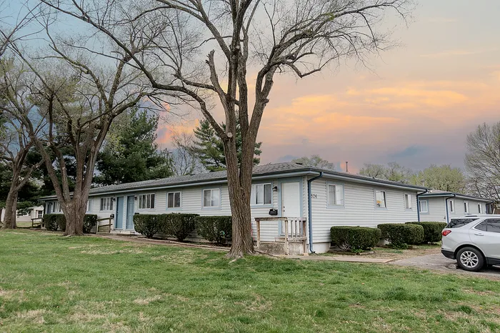 Wide-angle exterior of a quadplex unit at Village Club Apartments in Springfield MO with mature trees, trimmed shrubs, and private front entrances facing a grassy lawn.