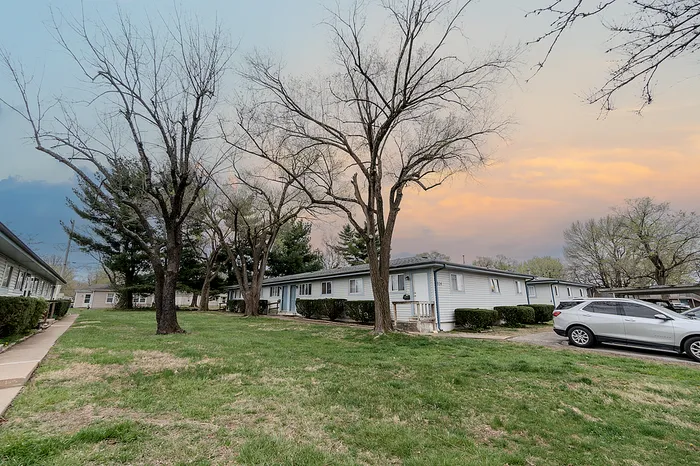 Village Club Apartments Springfield MO community layout featuring multiple one-story buildings with a central grassy courtyard and tall trees lining the sidewalk under a colorful evening sky.