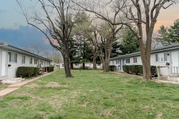 Community view at Village Club Apartments Springfield MO with multiple quadplex buildings, green space between units, and a paved parking area with vehicles near unit entrances.