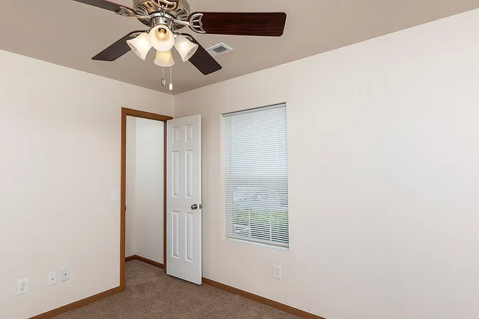 Carpeted bedroom at Lakewood Village Apartments in Springfield, MO, featuring a ceiling fan with light fixture, a window with blinds for natural light, and an open door leading to the hallway.