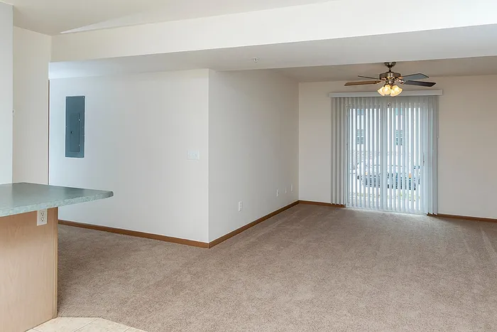 Open floor plan showcasing plush carpeted living space with a ceiling fan and large sliding glass door that leads to a private patio area, letting in plenty of natural light.