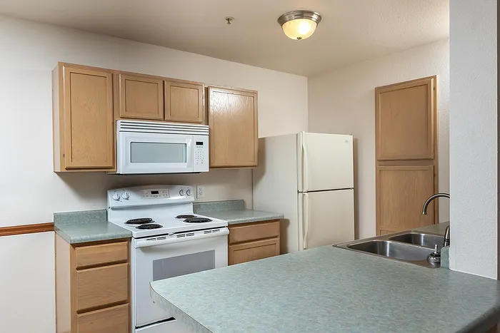 Functional kitchen with light wood cabinetry, white appliances including a microwave and stove, and a wraparound countertop with double sink&mdash;perfect for meal prep or casual dining.
