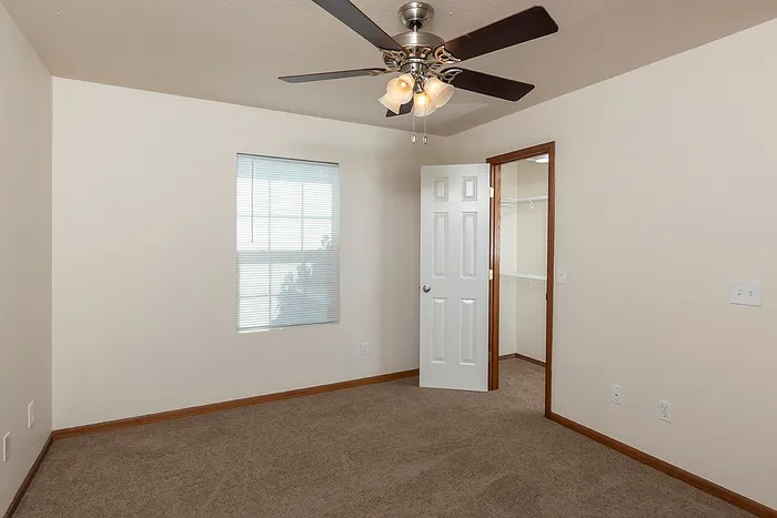 Bedroom with beige carpet, a ceiling fan with light fixture, and a large window with blinds. The open door leads to a walk-in closet with white wire shelving in this 2-bedroom, 2-bath Lakewood Village apartment.