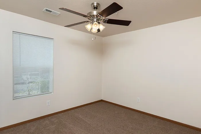 Bright and neutral-toned bedroom at Lakewood Village Apartments in Springfield, MO, featuring soft carpet, a ceiling fan with lights, and a window with blinds offering natural light and privacy.