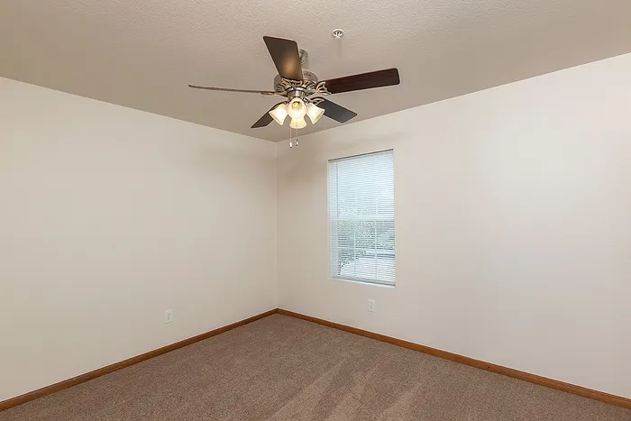 Spacious bedroom at Lakewood Village Apartments in Springfield, MO, featuring neutral carpeting, a large window with blinds, and a ceiling fan with light fixture&mdash;ideal for comfortable and quiet living.