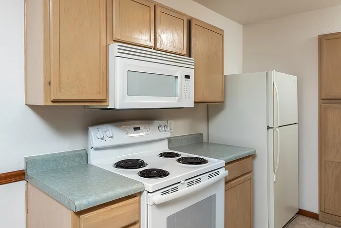 Close-up view of the kitchen at Lakewood Village Apartments in Springfield, MO, featuring a white electric stove with coil burners, built-in microwave, refrigerator, and light oak cabinetry with laminate countertops.