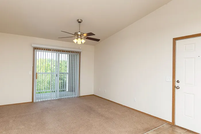 Comfortable living space with plush carpeting, ceiling fan, and vertical blinds that open to a private balcony, perfect for enjoying the outdoors.