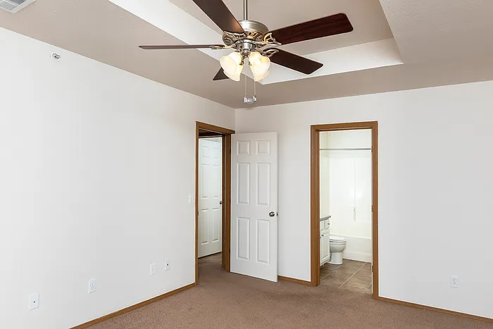 Spacious primary bedroom at Lakewood Village featuring neutral-tone carpet, tray ceiling with ceiling fan, and direct access to an en-suite bathroom with tile flooring.