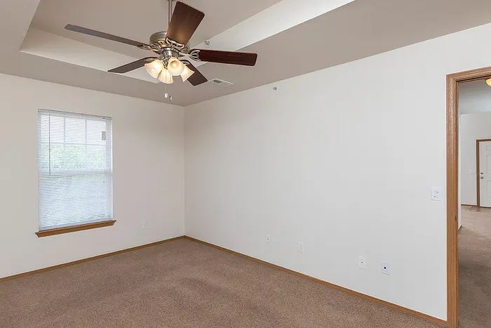 Spacious bedroom in a three-bedroom apartment at Lakewood Village, featuring plush carpeting, neutral walls, a large window with blinds, and a ceiling fan with overhead lighting.