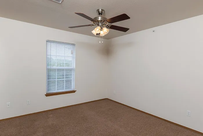 Another view of the Lakewood Village apartment bedroom showcasing a wide window, warm carpeting, and ceiling fan, offering a comfortable and bright living space.