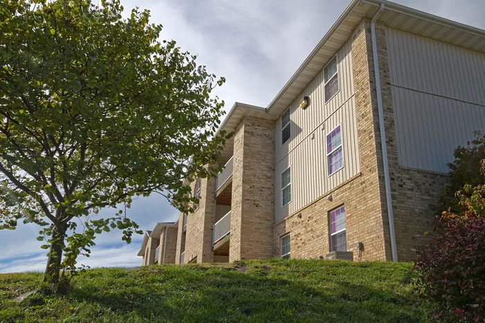 Exterior view of Lakewood Village Apartments in Springfield, Missouri, showcasing a multi-story brick and siding building with private balconies, mature trees, and a grassy hillside under a bright sky.