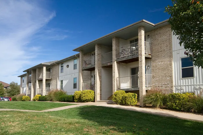 Front-facing view of Lakewood Village Apartments in Springfield, MO, featuring light brick and white panel exteriors, private balconies, manicured landscaping, and a clear blue sky&mdash;highlighting a peaceful and well-maintained apartment community.