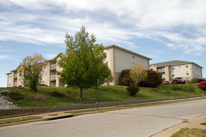 Wide street view of Lakewood Village Apartments in Springfield, Missouri, featuring neutral-toned brick buildings, mature trees, and well-maintained landscaping&mdash;ideal for peaceful apartment living near local conveniences.