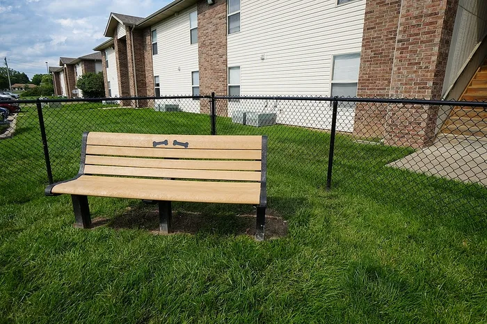 A wooden bench with dog bone cutouts sits on a grassy lawn inside a black chain-link fence at Huntley Ridge East Apartments in Springfield, MO. Brick and white siding apartment buildings are visible in the background.